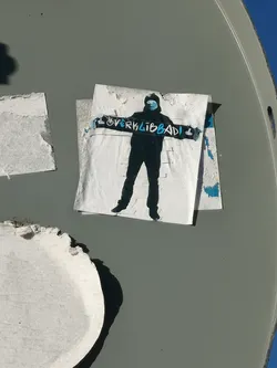A monochrome sticker featuring a stylized, dark silhouette of a person standing, possibly wearing a hoodie or beanie. The figure's face is partially obscured by a light blue face mask. They hold a long, horizontal banner or scarf across their chest, which is dark with light blue text that reads 'OVERKLIBBAD!' and a small symbol resembling a fist or hand gesture at the right end. The sticker itself is white, showing signs of wear and peeling at the edges, and is affixed to a grey, curved surface.