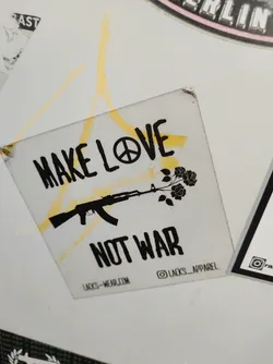A sticker with a pacifist message. It features an AK-47 rifle with roses growing out of the barrel, symbolizing the transformation from violence to peace. The text 'Make Love Not War' is prominently displayed, along with a peace symbol.
