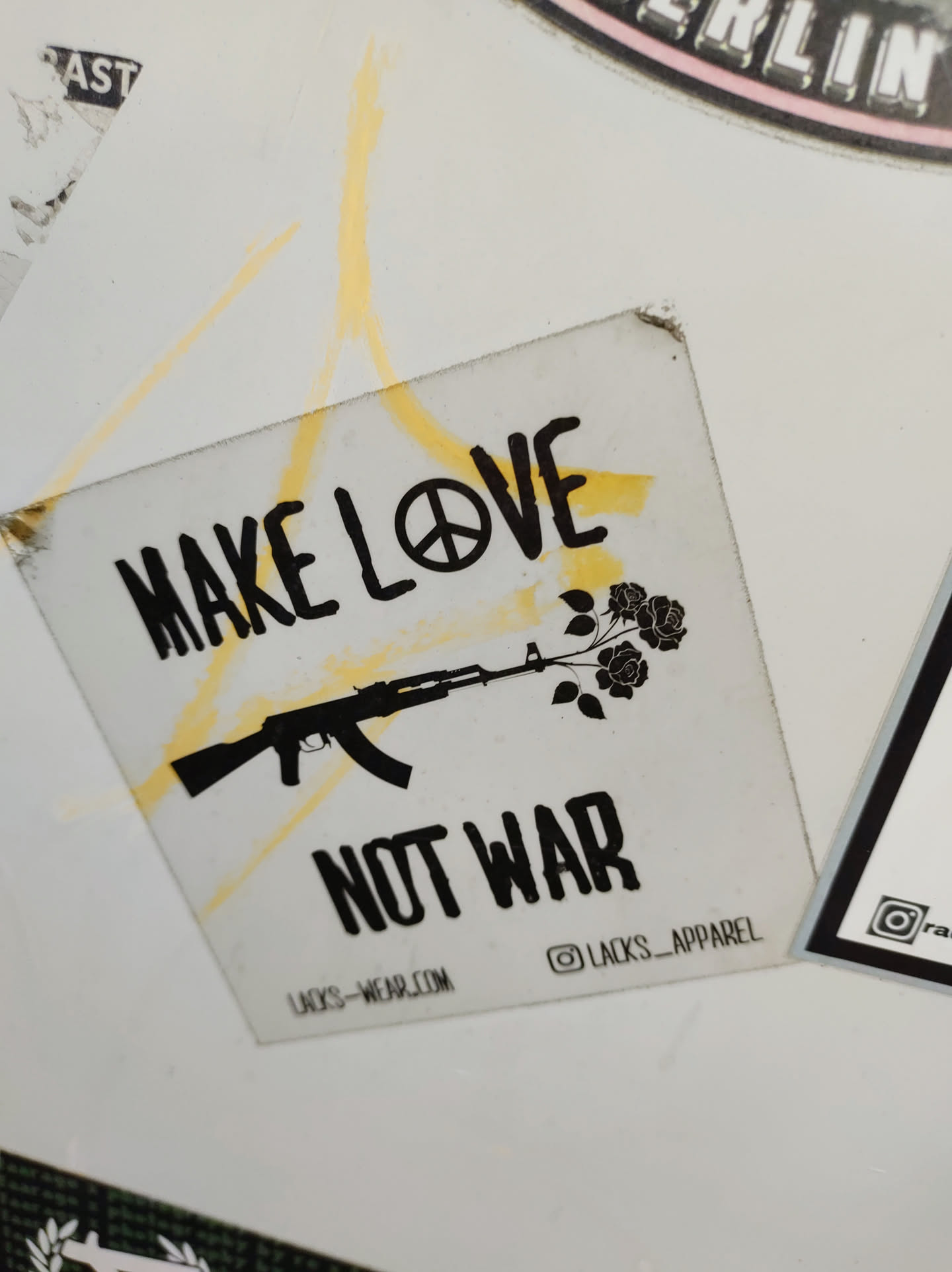 A sticker with a pacifist message. It features an AK-47 rifle with roses growing out of the barrel, symbolizing the transformation from violence to peace. The text 'Make Love Not War' is prominently displayed, along with a peace symbol.