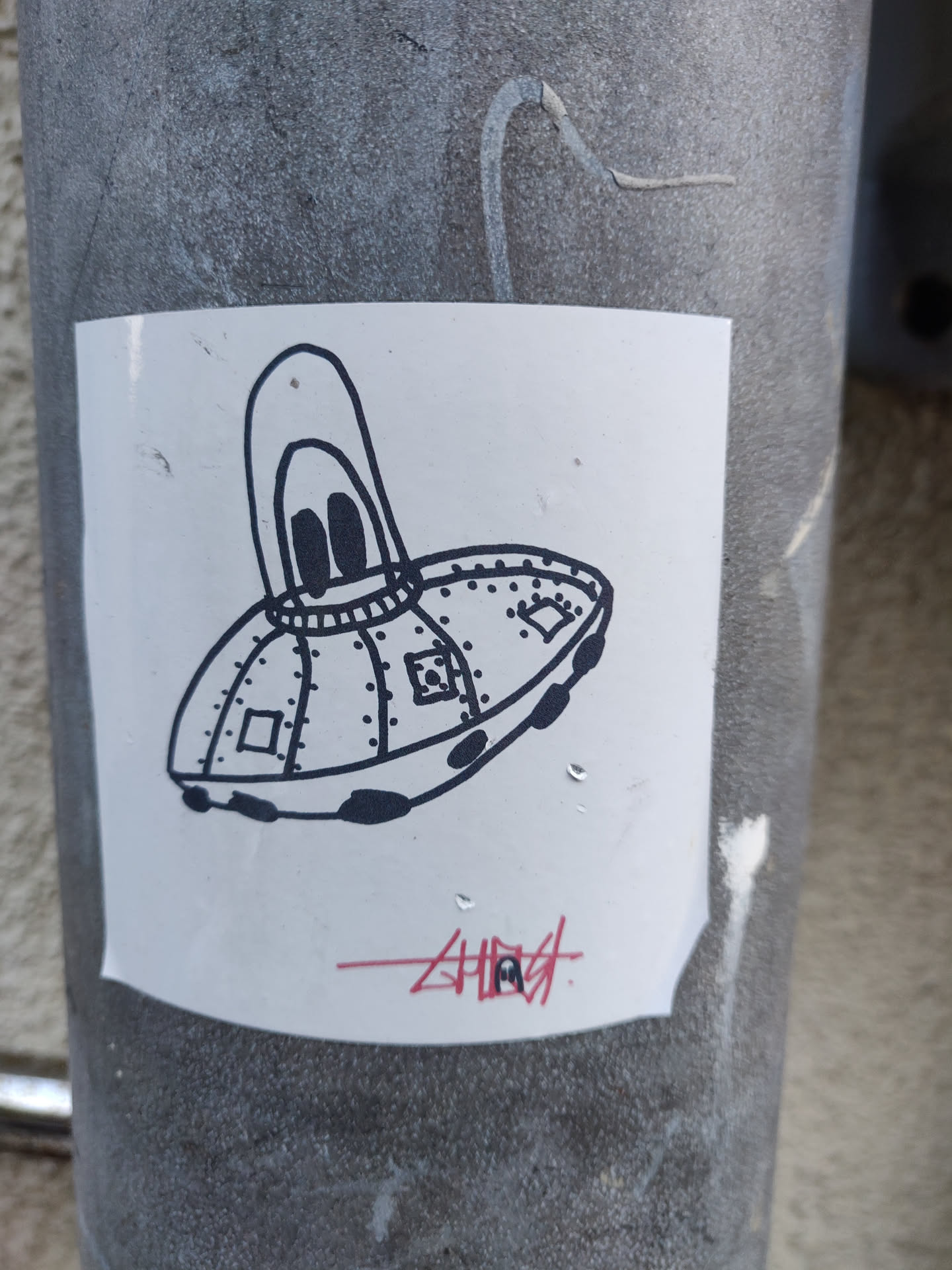 A white sticker with a black and white illustration of a cartoon UFO. The UFO has a rounded top with two small circles that look like eyes and rivets along its body. The sticker has the word "Ghost" written underneath in red.