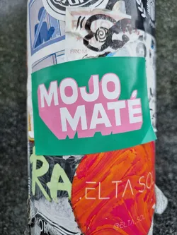 A green and pink sticker with the words "MOJO MATÉ" in a stylized font. The sticker is attached to a surface covered in other stickers.