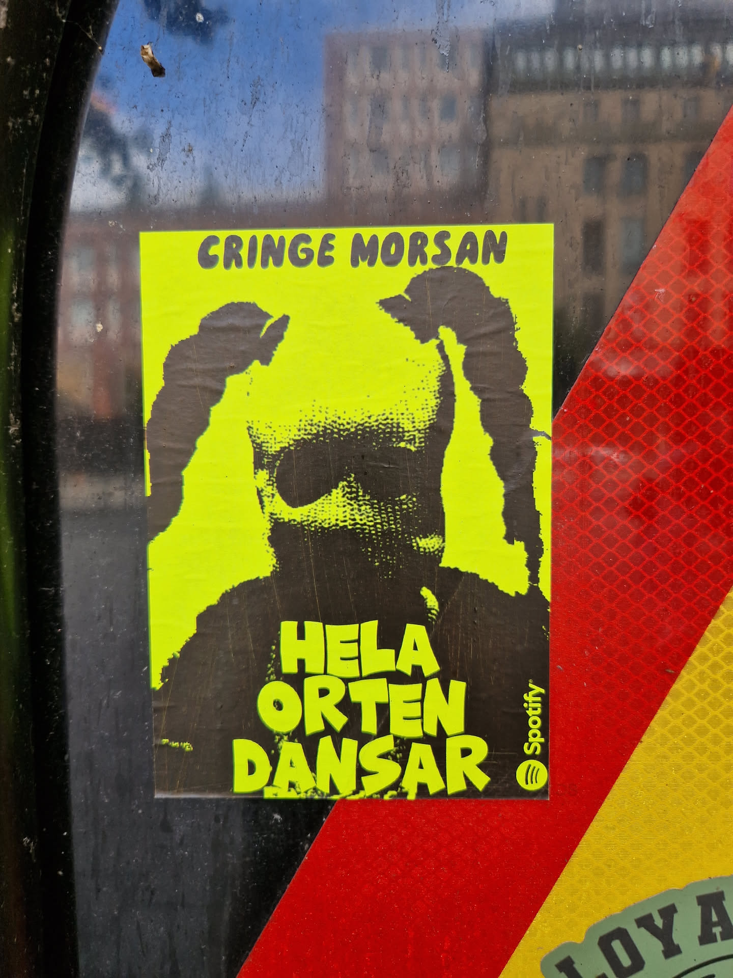 A yellow and black sticker featuring an illustration of a person with their face covered, with the text "Cringe Morsan" at the top and "Hela Orten Dansar" at the bottom.  There is a small Spotify logo in the bottom right corner.