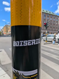 A "BOISERIE" sticker is adhered to a pole with a yellow reflective top. The sticker is white with a black outline, and the text is in a slightly futuristic, blocky font.