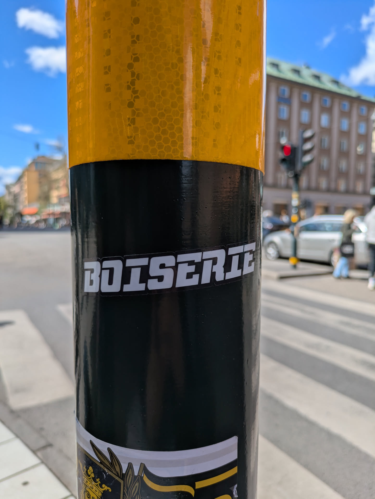 A "BOISERIE" sticker is adhered to a pole with a yellow reflective top. The sticker is white with a black outline, and the text is in a slightly futuristic, blocky font.