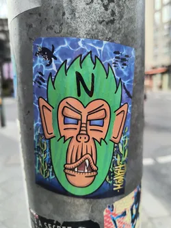 A sticker featuring a cartoon monkey head with green spiky hair, set against a blue underwater background with fish and turtles. The monkey has a grumpy expression and prominent teeth. The letter N is prominently displayed in the monkey's hair.  The artist's signature is visible in the bottom right corner.