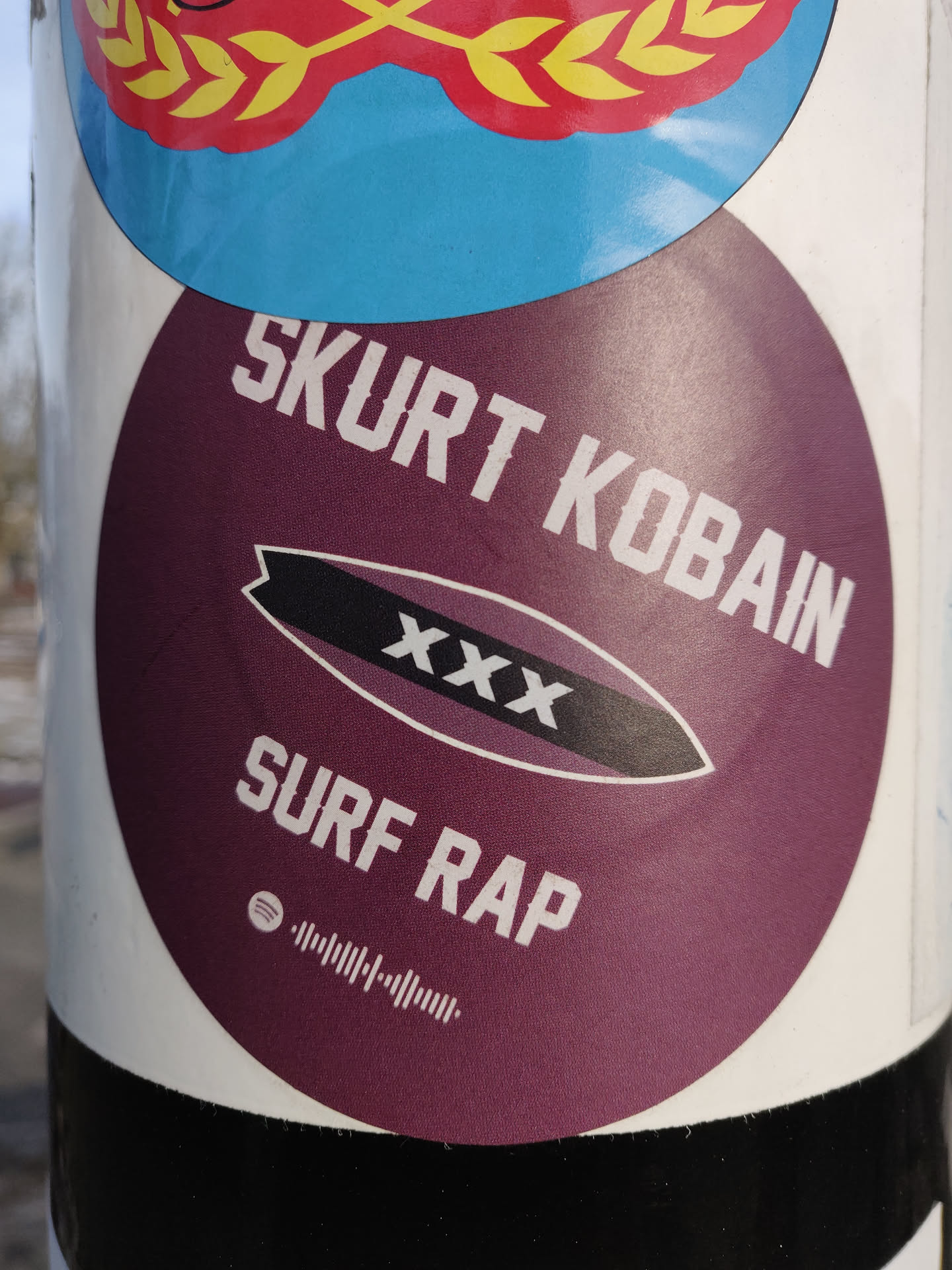 Round maroon sticker with the text "SKURT KOBAIN" on top, a surfboard graphic with "XXX" on it in the middle, and "SURF RAP" at the bottom. There is also a Spotify code at the bottom.
