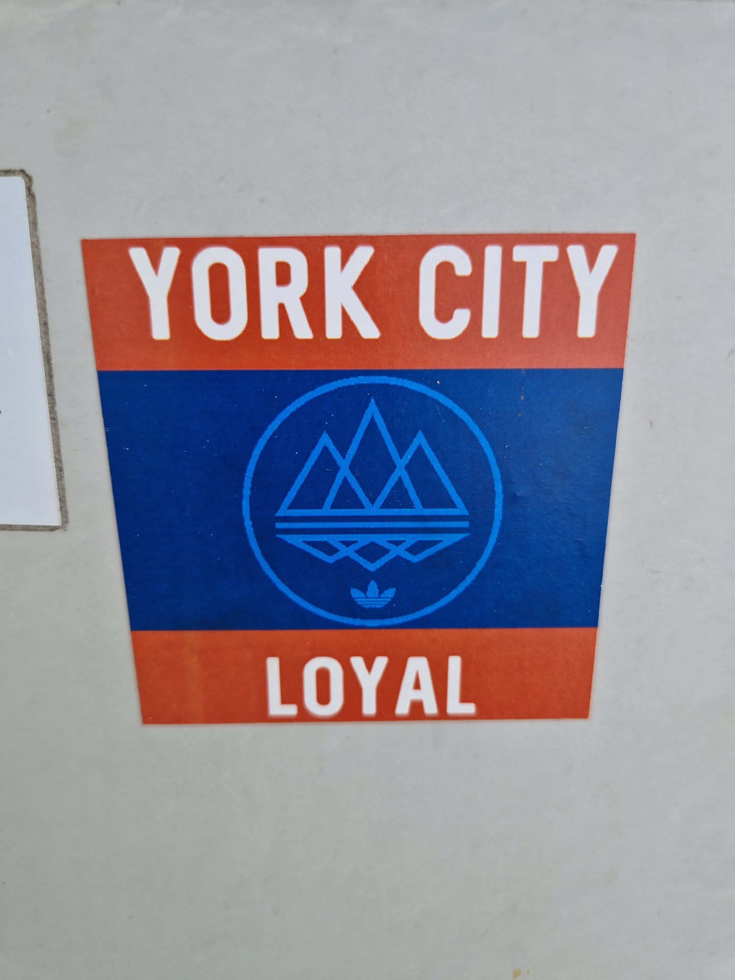 A sticker featuring the text "YORK CITY" at the top, a logo with three stylized mountains and the Adidas logo in the center, and the text "LOYAL" at the bottom. The colors used are blue, orange and white.