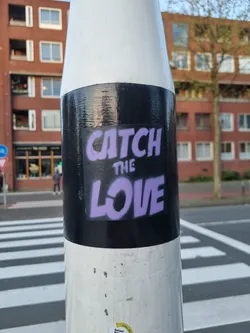 A purple and black sticker on a white pole with the text "CATCH THE LOVE". The sticker is placed on a black background.