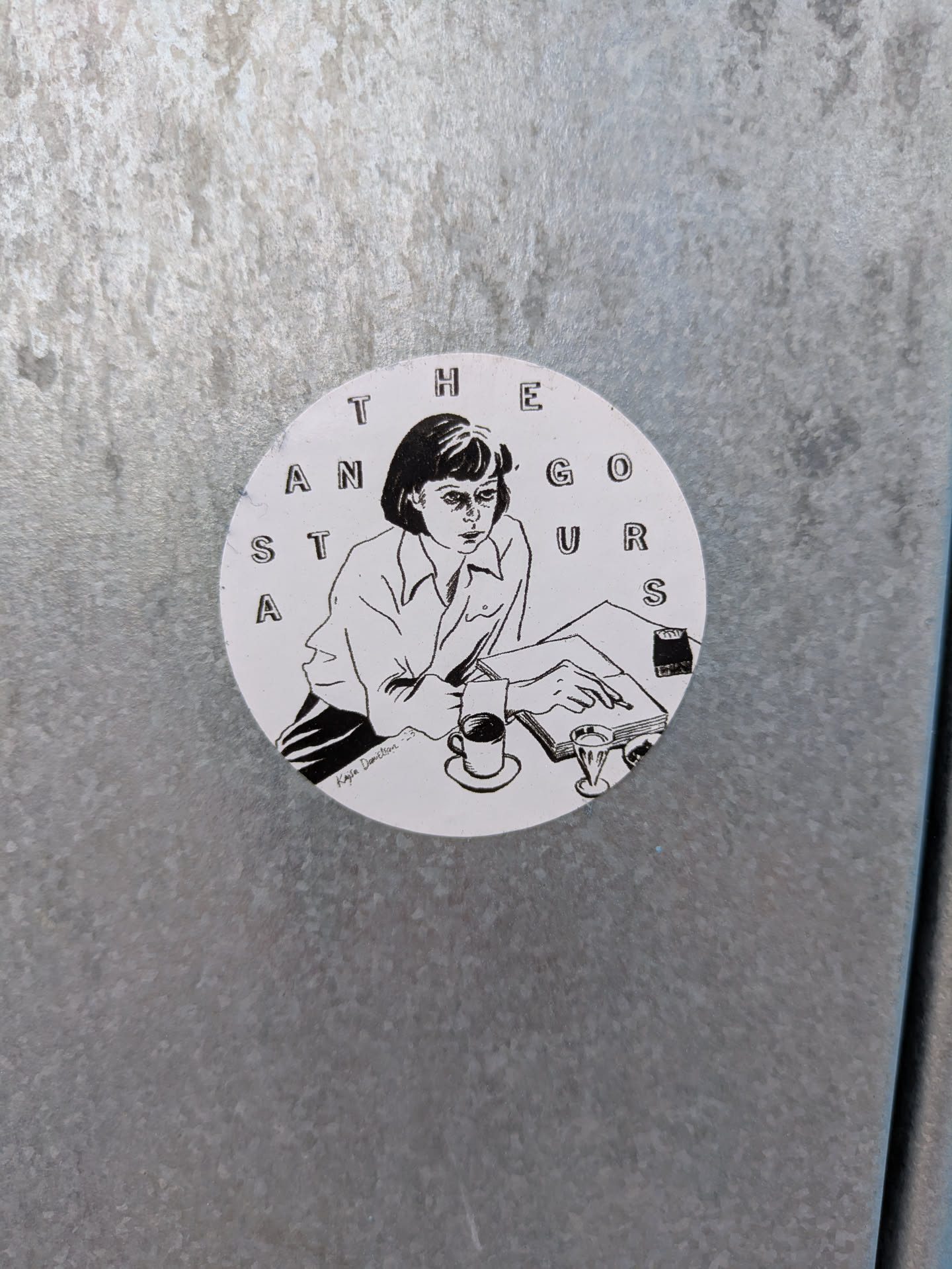 The sticker is circular and features a black and white illustration of a young woman with short hair, wearing a collared shirt and striped pants. She is seated at a table, with her head tilted down as she reads a book or document. Her left hand rests on the page, holding a pen. To her right, there is a cup of coffee on a saucer and a small glass. The text 'THE GURUS' is arranged in a circular pattern above her, with some letters incomplete. Below her, the text 'Kajsa Danielsson' is written in a cursive font.