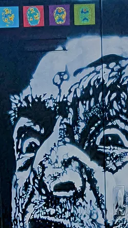 The image features a close-up view of a sticker art piece. At the top are small square stickers in different colors: blue, red, purple, and green. Each one contains abstract shapes, possibly representing a brain or some other organ. Below these is a larger artwork depicting a face. The face is rendered in an abstract style, with the contours and features created by blotches of dark and light shades, resembling an almost skeletal or decayed appearance. The style suggests a stencil or spray-painted technique, adding to the gritty urban aesthetic. The face's features are still somewhat discernible, with indications of eyes, nose, and mouth.
