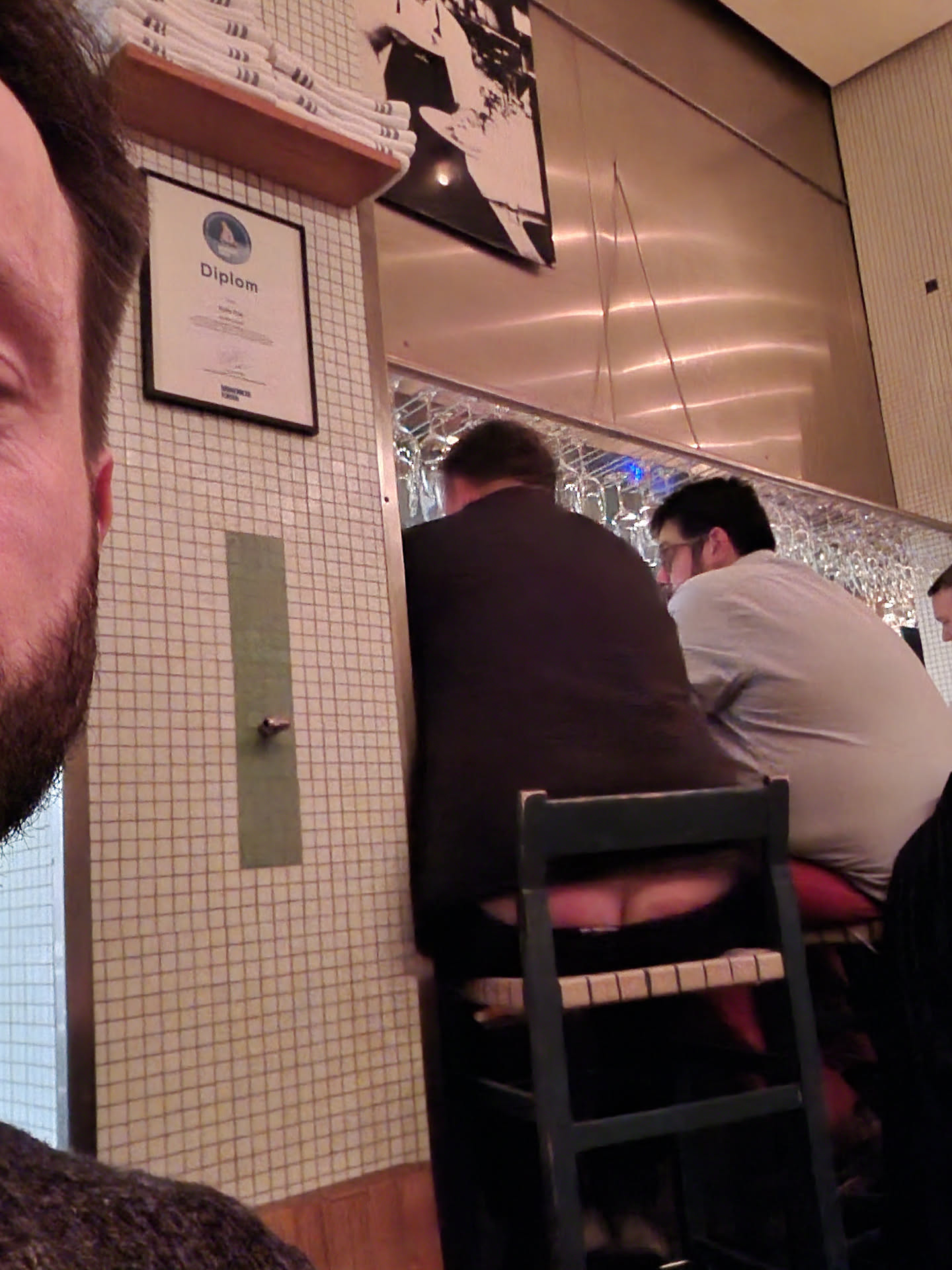 This image shows the back of a person sitting on a bar stool.  The focus is primarily on the person's rear and the stool itself. The background includes other bar patrons and a tiled wall.
