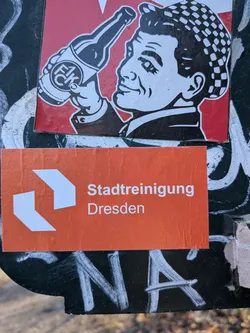 A rectangular orange sticker featuring two white, angular arrow-like shapes on the left side, pointing towards the right. To their right, in white sans-serif text, are the words 'Stadtreinigung' on the upper line and 'Dresden' on the lower line. The sticker has a slightly textured appearance and minor creases, suggesting it is a physical adhesive label.