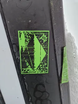 A lime green rectangular sticker stands out against a dark surface.  It features a geometric, possibly abstract, design in black. The letters "ZRA" are visible near the bottom. The sticker has a slightly distressed or textured look, as if printed on slightly rough paper. 