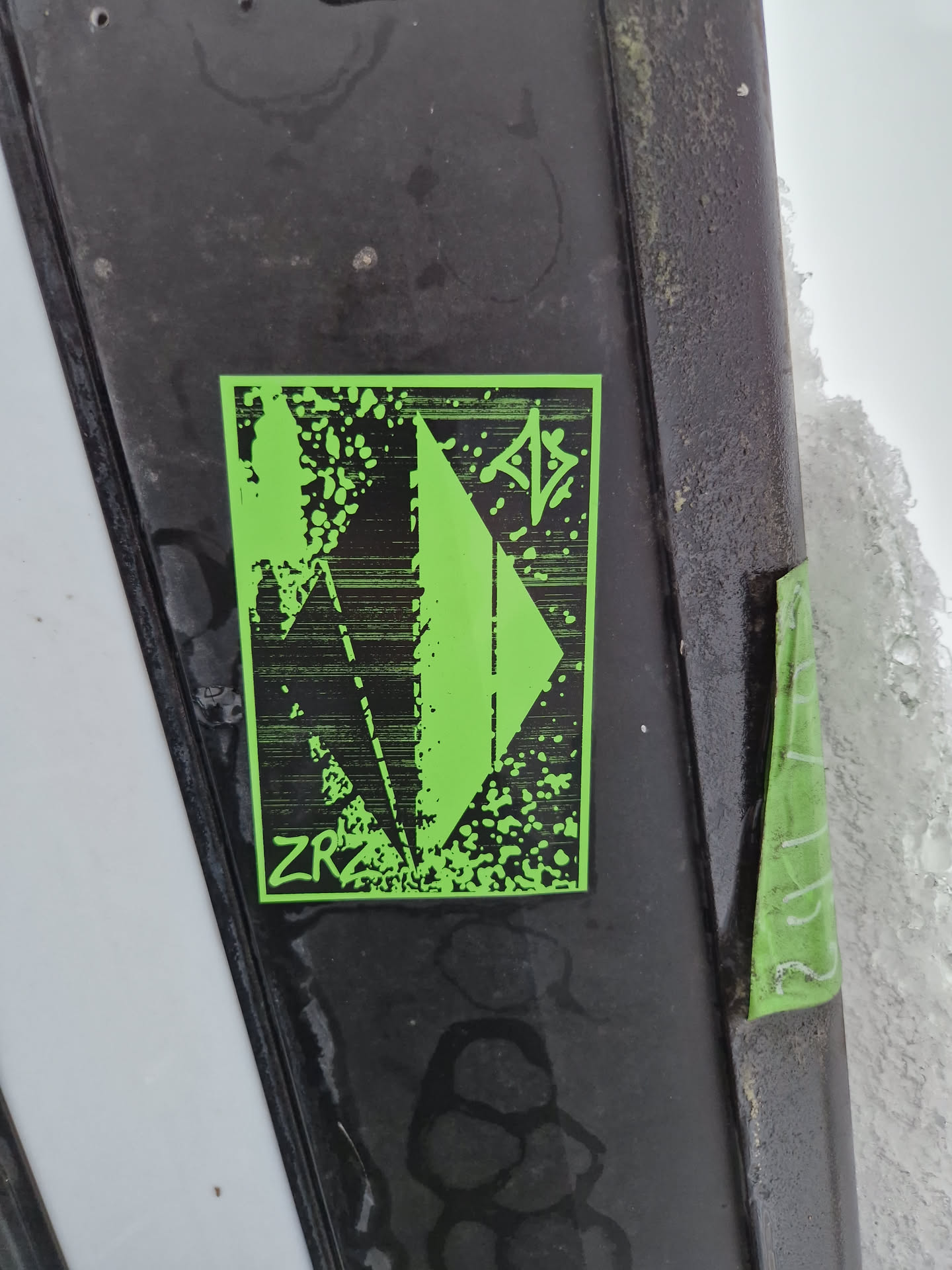 A lime green rectangular sticker stands out against a dark surface.  It features a geometric, possibly abstract, design in black. The letters "ZRA" are visible near the bottom. The sticker has a slightly distressed or textured look, as if printed on slightly rough paper. 