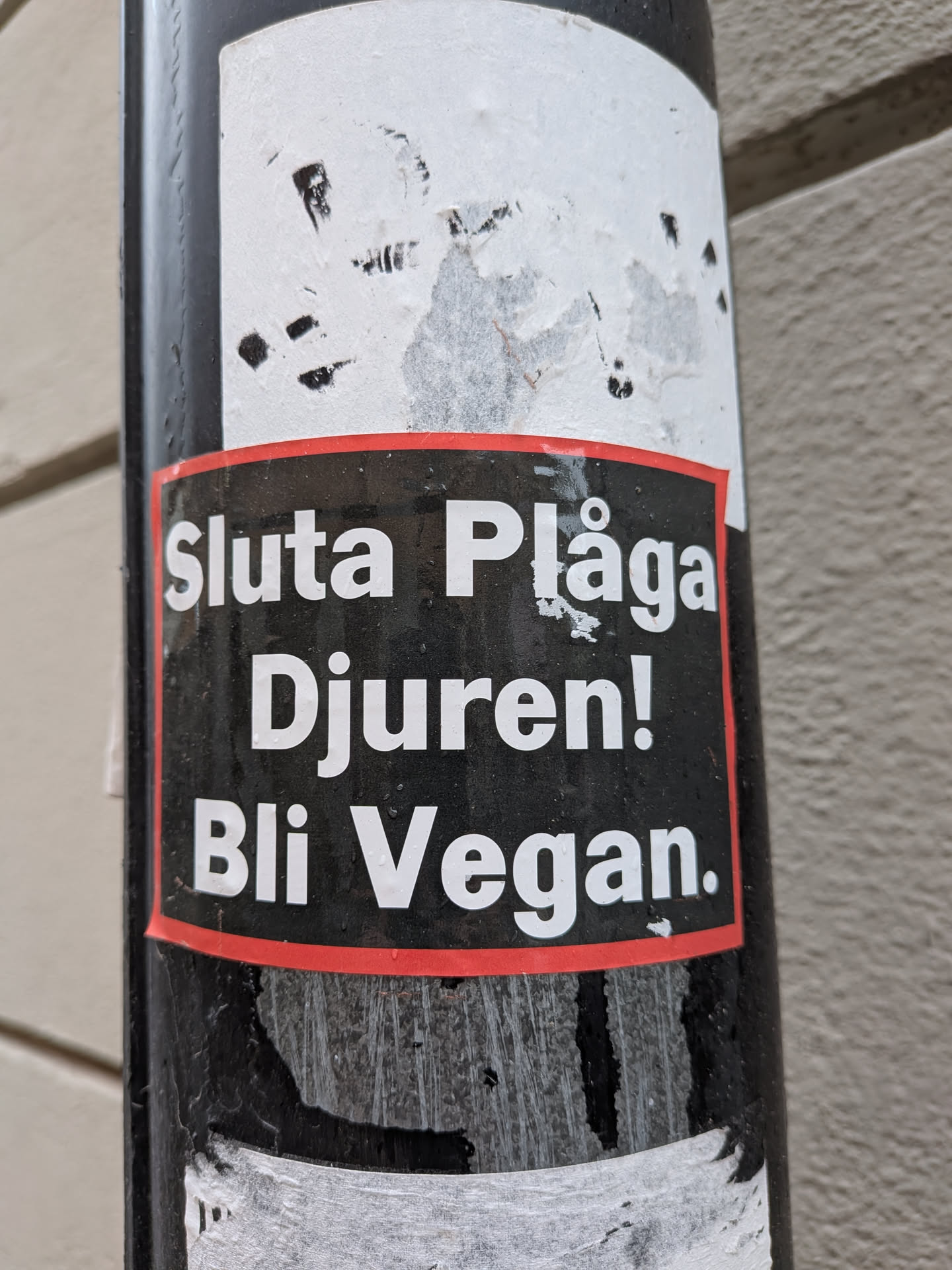 A rectangular sticker with a black background and a thin red border. The text is prominently displayed in white, sans-serif font, spread across three lines. The sticker appears to be affixed to a dark, possibly metallic, pole, showing some signs of wear and tear, particularly around its edges and the top where some of the black background is chipped away, revealing the underlying surface or adhesive.