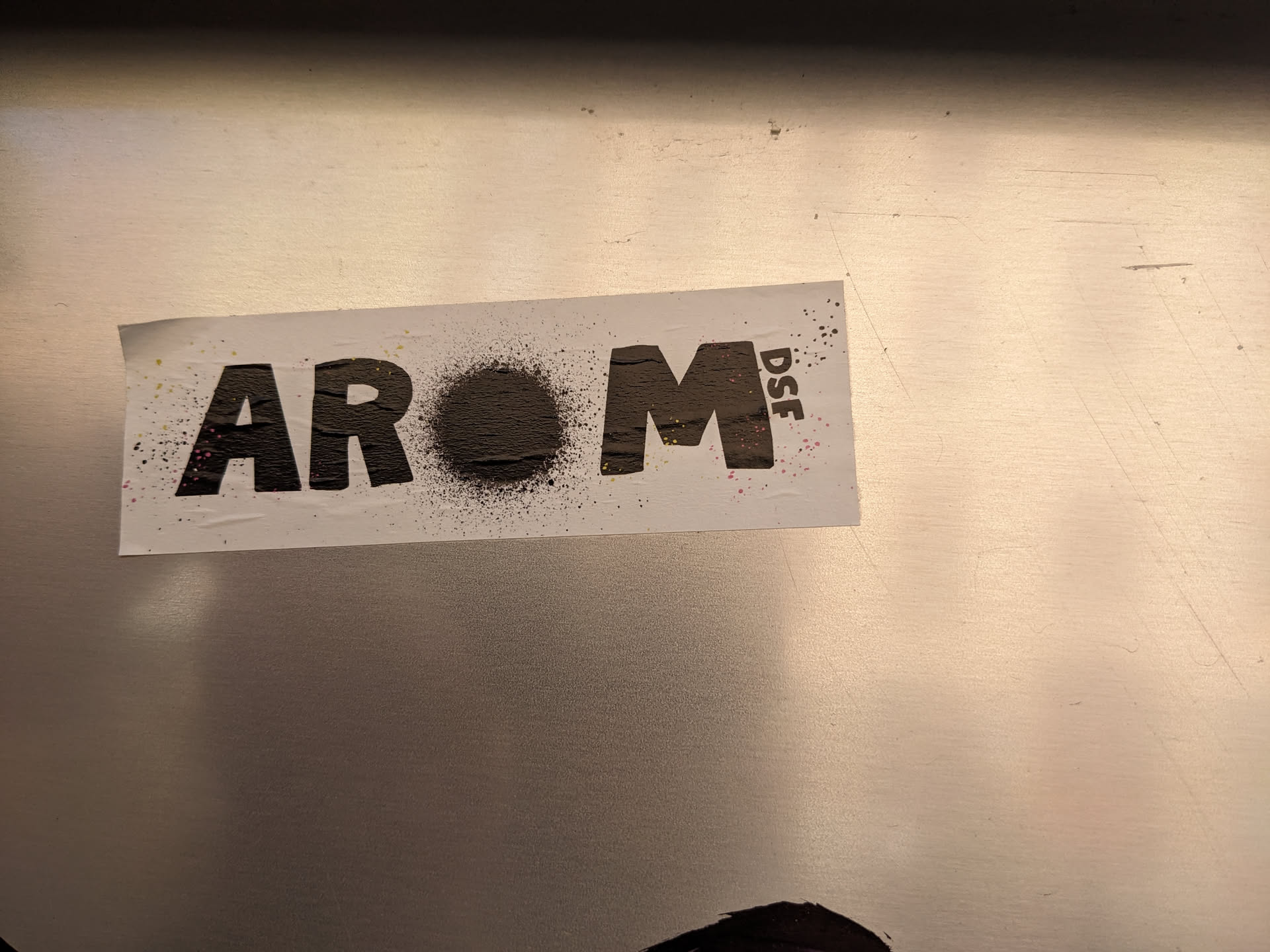Close-up shot of a sticker featuring the word AROM in stylized lettering, interspersed with a small circular graphic.  The overall aesthetic is minimalist and slightly gritty, suggesting a street art or urban style.  The small text "DSF" adds another layer of possible meaning or branding.