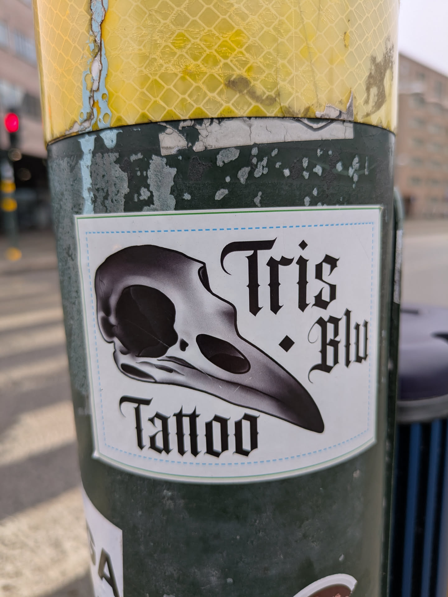 A sticker on a pole advertises 'Tris Blu Tattoo'. The sticker features a stylized raven skull design.