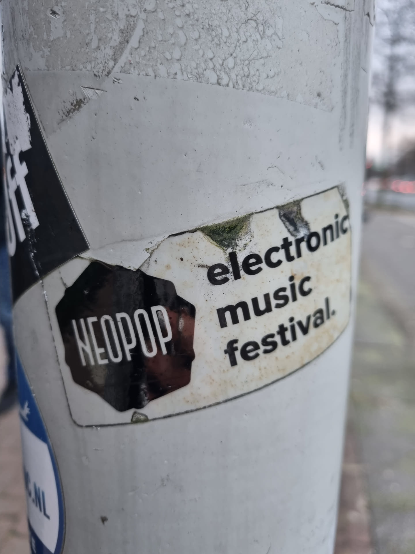 A sticker advertising the Neopop Electronic Music Festival. The sticker is black and white and features the festival's logo and name.  The sticker is slightly damaged and dirty from being affixed to an outdoor surface.