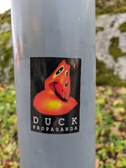 A sticker featuring a stylized orange duck-like figure with the text 'DUCK PROPAGANDA' underneath. The sticker is affixed to a gray pole outdoors.