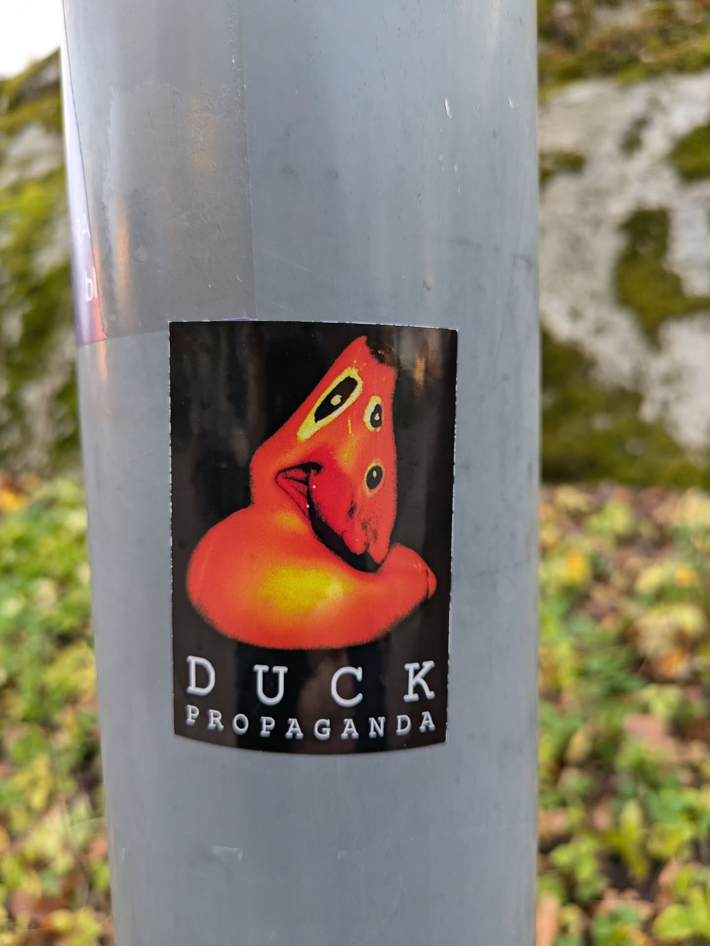 A sticker featuring a stylized orange duck-like figure with the text 'DUCK PROPAGANDA' underneath. The sticker is affixed to a gray pole outdoors.