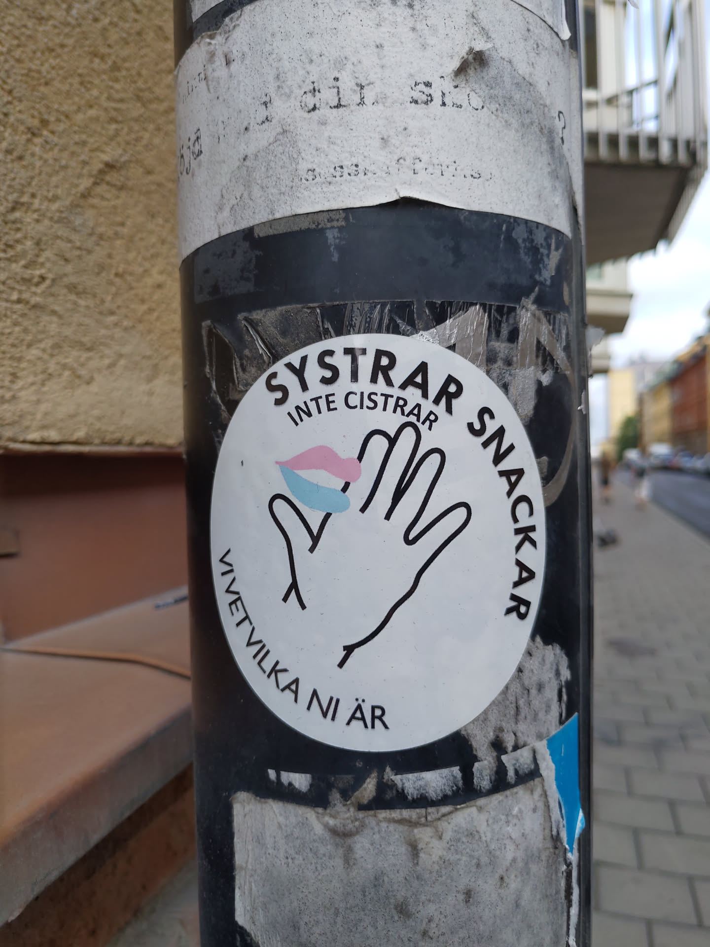 A white round sticker with black text and a graphic of a hand with a small pink, blue and white lips design. The text on the sticker says "SYSTRAR INTE CISTRAR SNACKAR" which translates to "Sisters don't silence, talk" and "VÄTVILKA NI ÄR" which translates to "who are you".