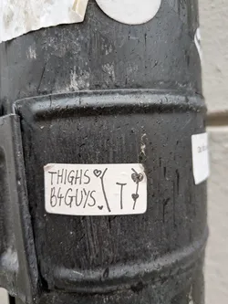 A rectangular, off-white sticker with slightly rounded corners, adhered to a dark, textured metal surface. The sticker features handwritten black text that reads 'THIGHS B4 GUYS'. A small black heart is drawn above the 'S' in 'THIGHS' and another below the 'S' in 'GUYS'. To the right of the main text, there is a larger, stylized black 'T' with two small black hearts drawn on it, one near the top and one near the bottom. The sticker appears slightly worn and has a homemade aesthetic.
