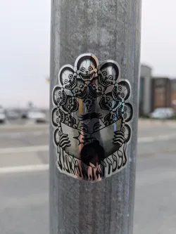 A silver holographic sticker affixed to a metal pole. The artwork is centered around a figure seemingly holding an open book. The figure is encircled by several smaller depictions of skulls, each with exaggerated grins. The style suggests dark fantasy or surrealism. No visible social media handles or QR codes are present.