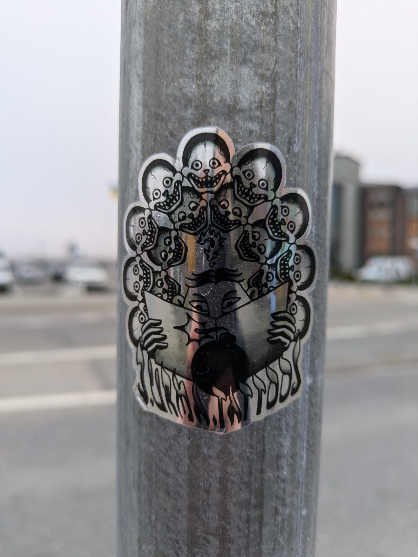 A silver holographic sticker affixed to a metal pole. The artwork is centered around a figure seemingly holding an open book. The figure is encircled by several smaller depictions of skulls, each with exaggerated grins. The style suggests dark fantasy or surrealism. No visible social media handles or QR codes are present.
