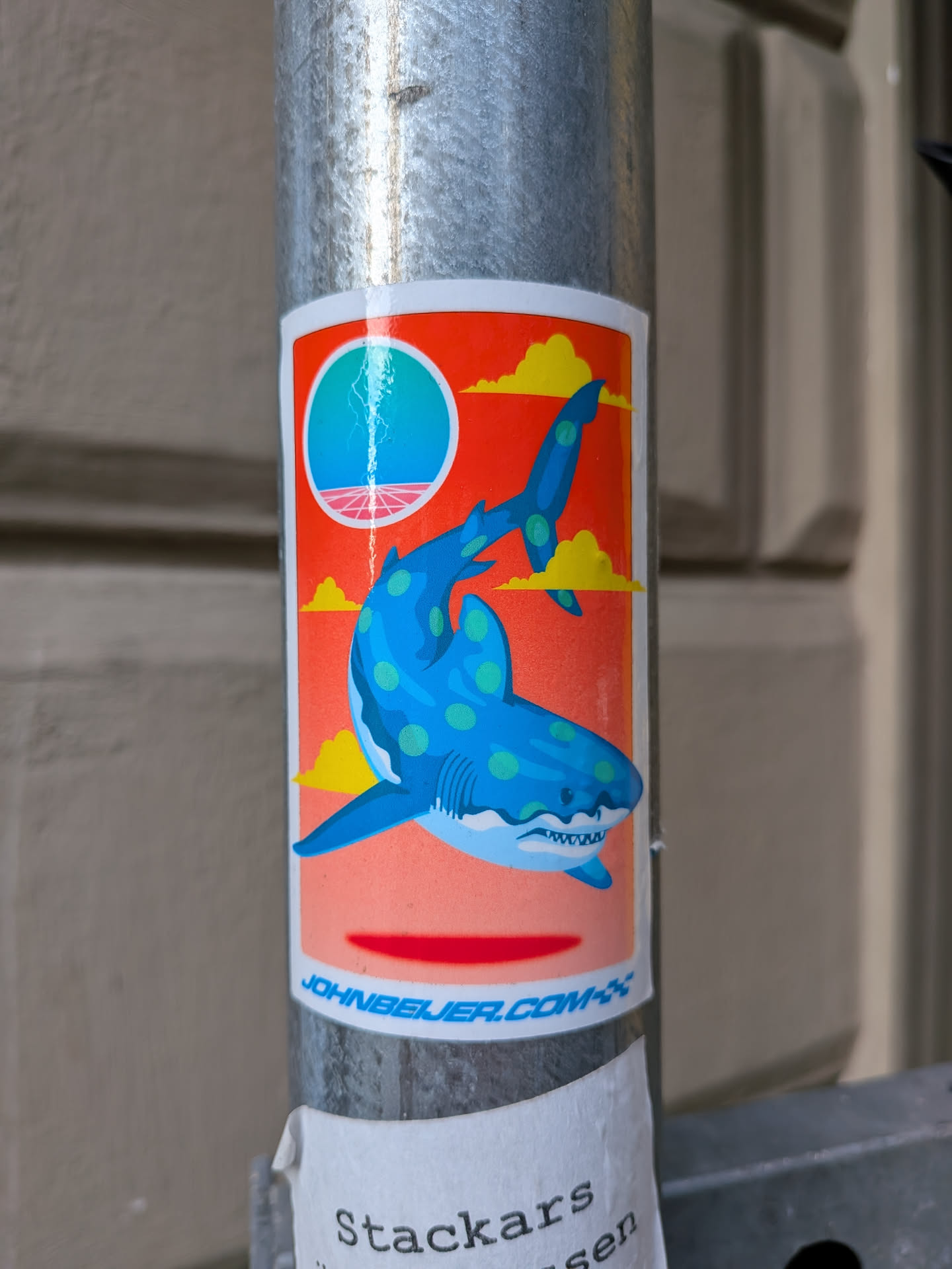 The sticker features a stylized blue shark with light green polka dots, swimming against a vibrant orange background. Above the shark, a circular motif depicts a blue orb with lightning striking from it, set against a pink horizon and stylized yellow clouds. The overall aesthetic has a retro-futuristic feel. The shark has its mouth open, revealing sharp white teeth. The bottom edge of the sticker has a white border with blue text.