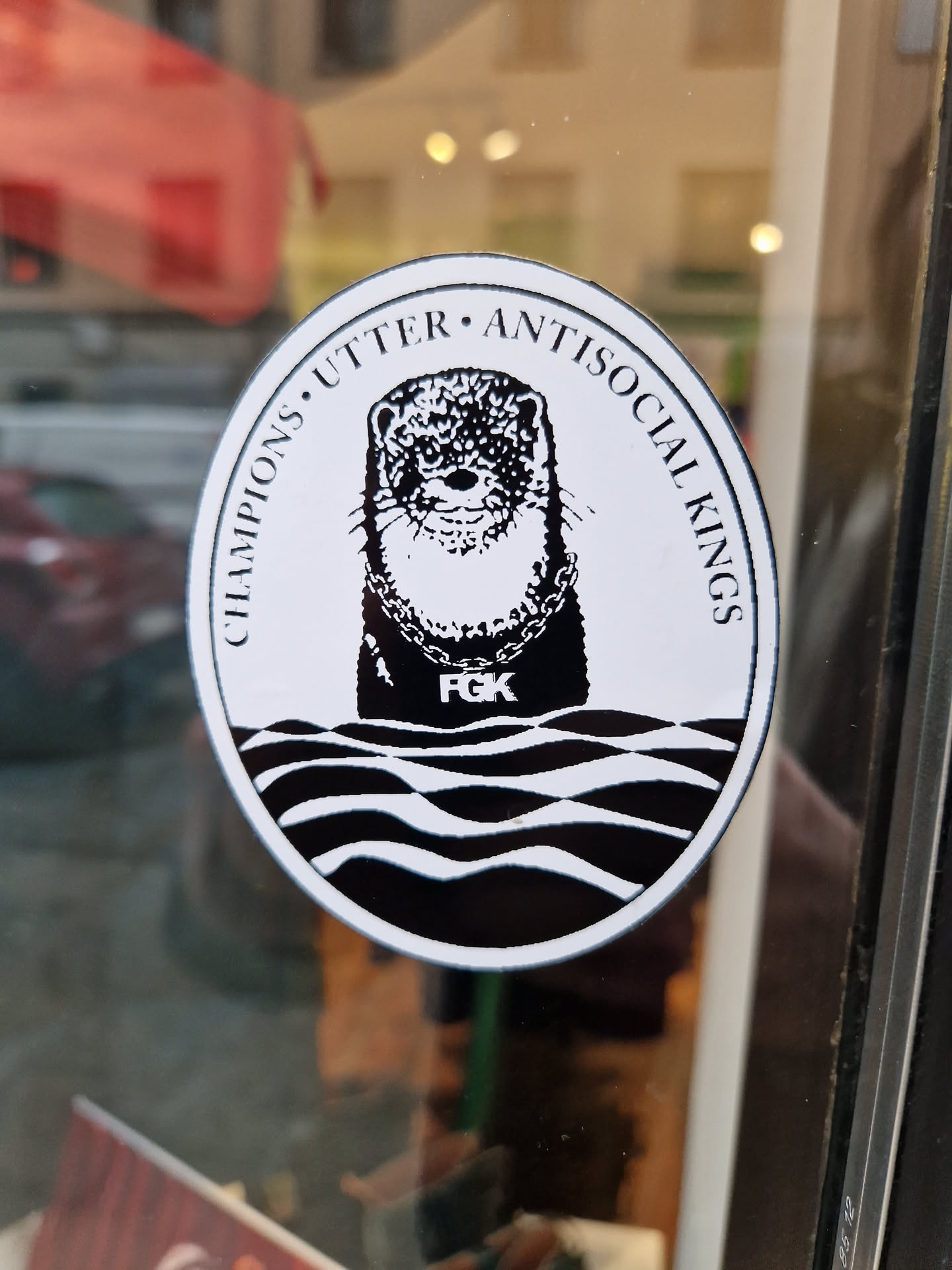 Black and white sticker of an otter wearing a gold chain with the text "Champions Utter Antisocial Kings" around it. The otter has the letters F.G.K on its chest. The sticker is oval-shaped and has a wave pattern at the bottom.