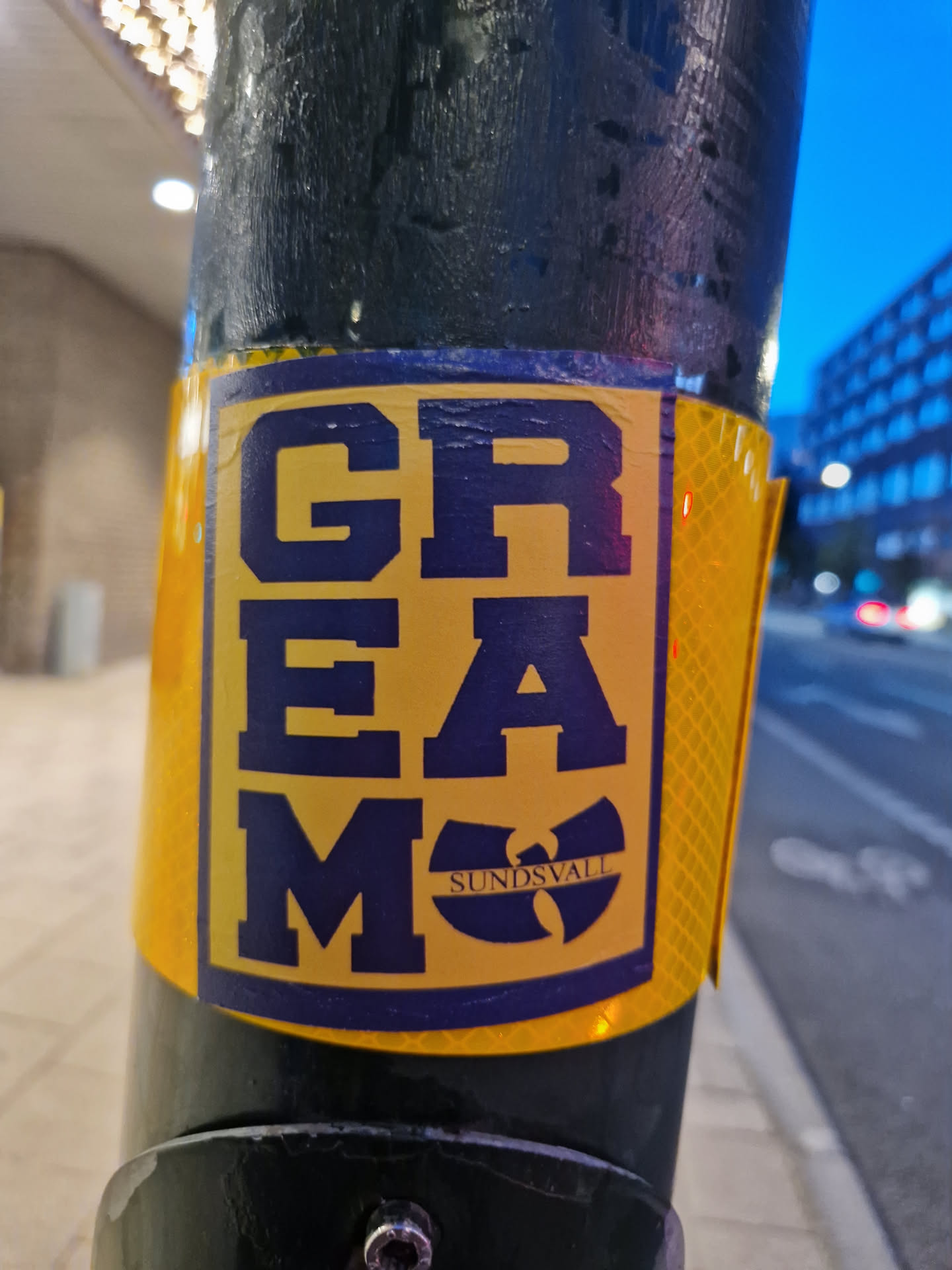A yellow and dark blue sticker featuring the stylized text 'GREMO' and the Wu-Tang Clan logo with 'SUNDSVALL' inscribed below it. The sticker is affixed to a dark pole outdoors.