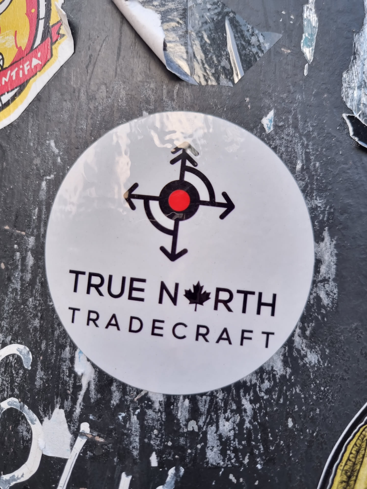 A white round sticker with a compass rose and the text "TRUE NORTH TRADERCRAFT". The design is simple and minimalist, with the text in a sans-serif font. The sticker is affixed to a dark-colored surface, which appears to be a wall or other outdoor structure. There are other stickers and graffiti visible in the background.