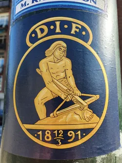 This sticker features a circular design with a dark blue background.  A gold figure, possibly representing a historical or mythological character, is depicted operating a large catapult.  The letters D.I.F. are inscribed above the figure. Below the figure, the numbers 1812 and 91 are visible, with a fraction (12/3) between them. The style is reminiscent of classic or vintage illustrations.