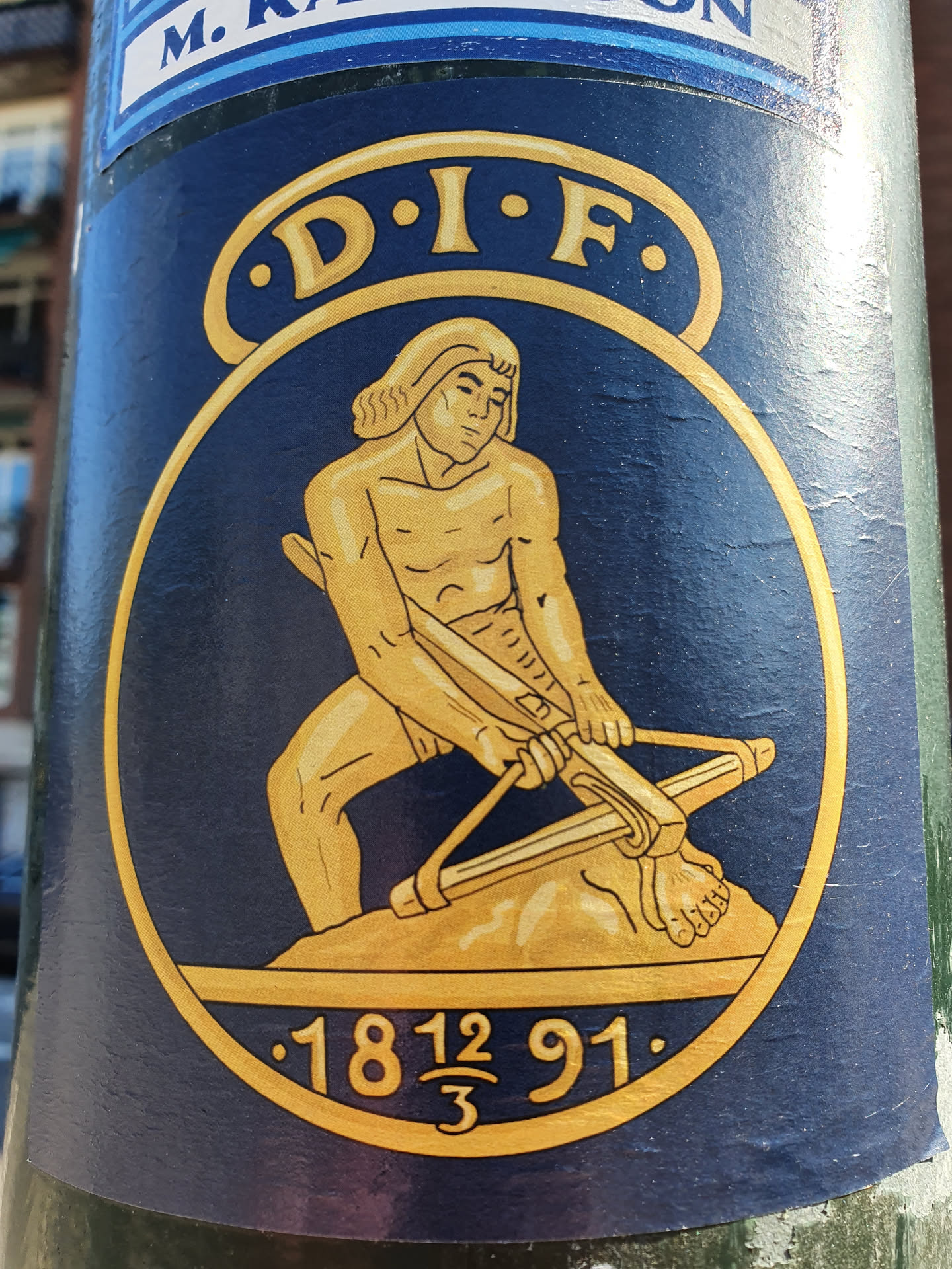 This sticker features a circular design with a dark blue background.  A gold figure, possibly representing a historical or mythological character, is depicted operating a large catapult.  The letters D.I.F. are inscribed above the figure. Below the figure, the numbers 1812 and 91 are visible, with a fraction (12/3) between them. The style is reminiscent of classic or vintage illustrations.