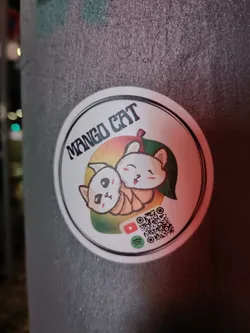 A round sticker featuring two cute cartoon cats nestled within a mango. The text "MANGO CAT" is prominently displayed at the top.  A QR code is included along with links to YouTube and Spotify.