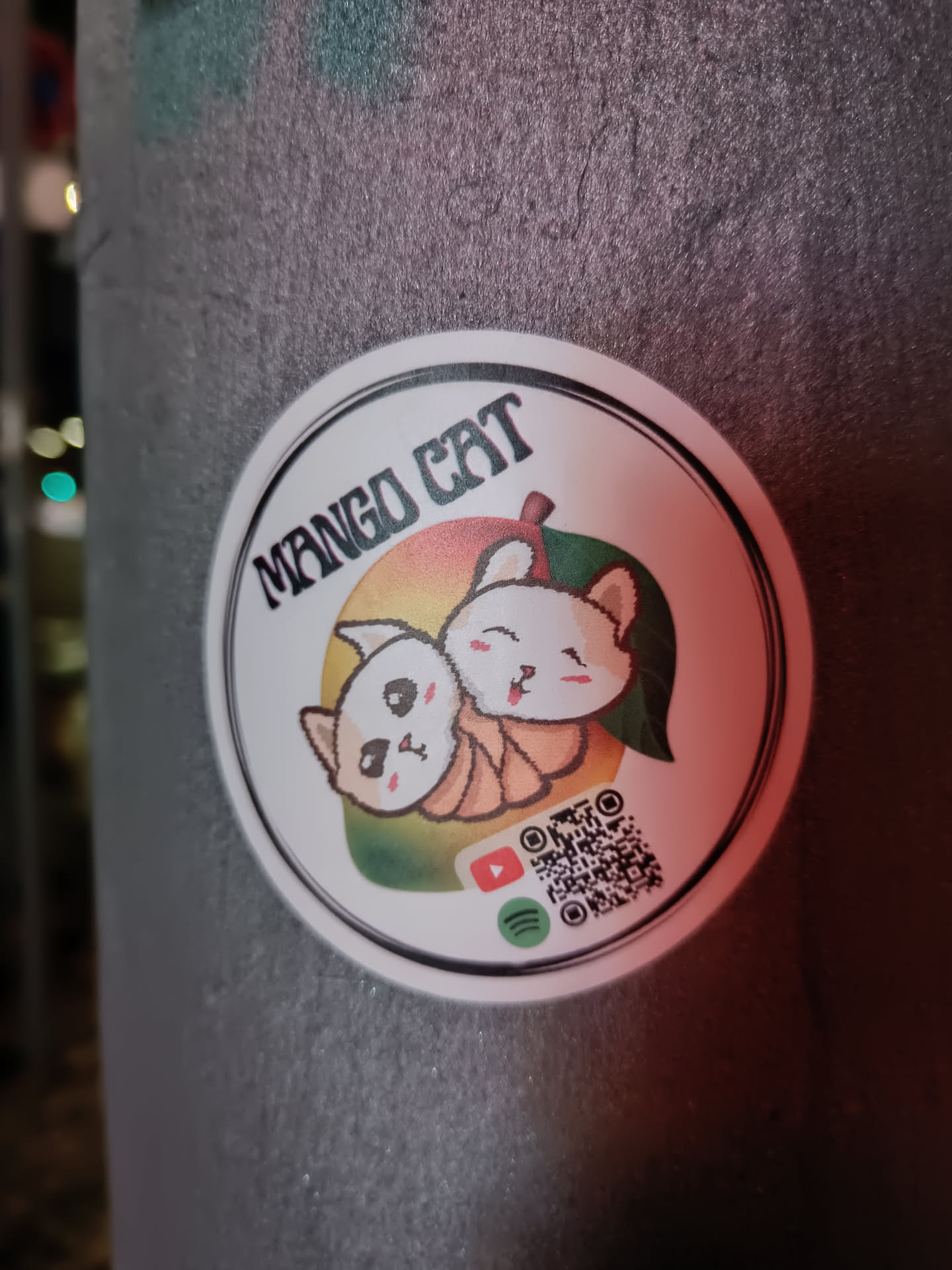 A round sticker featuring two cute cartoon cats nestled within a mango. The text "MANGO CAT" is prominently displayed at the top.  A QR code is included along with links to YouTube and Spotify.