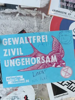 A light blue sticker with a bird and the text 'Gewaltfrei zivil ungehorsam' (non-violent civil disobedience) and the Extinction Rebellion logo.  There's also some additional handwritten text on the sticker.