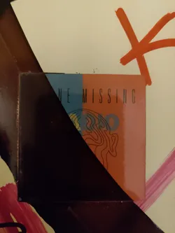 A sticker featuring a minimalist design split into two colors, dark blue and light orange. The text 'ONE MISSING' is visible.  A stylized brain-like image is partially visible on the orange half. The sticker is affixed to a surface with other painted markings.
