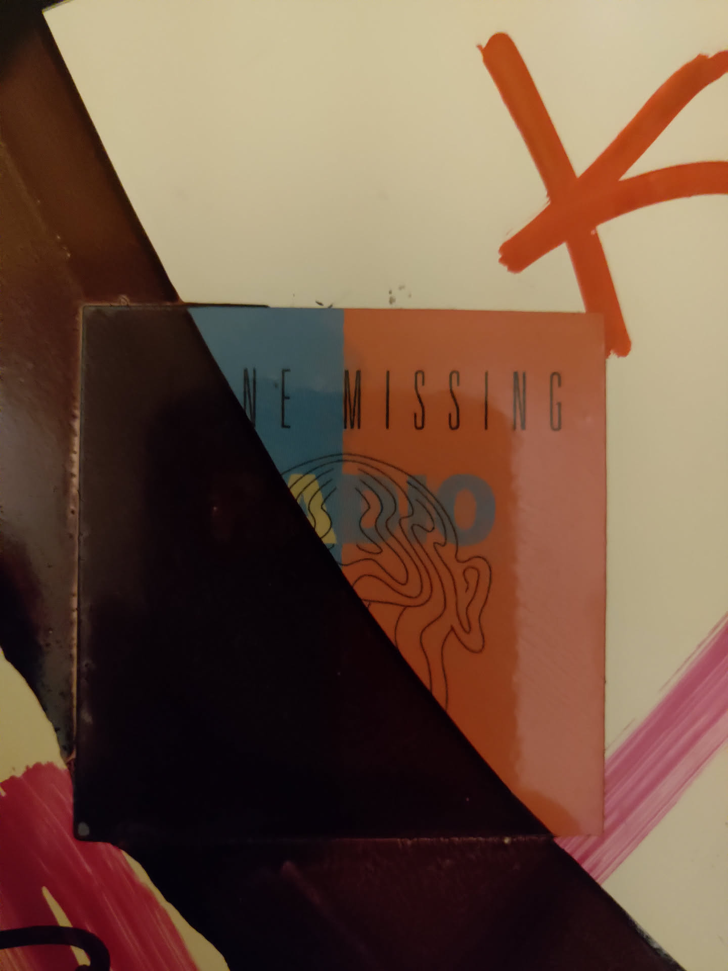 A sticker featuring a minimalist design split into two colors, dark blue and light orange. The text 'ONE MISSING' is visible.  A stylized brain-like image is partially visible on the orange half. The sticker is affixed to a surface with other painted markings.