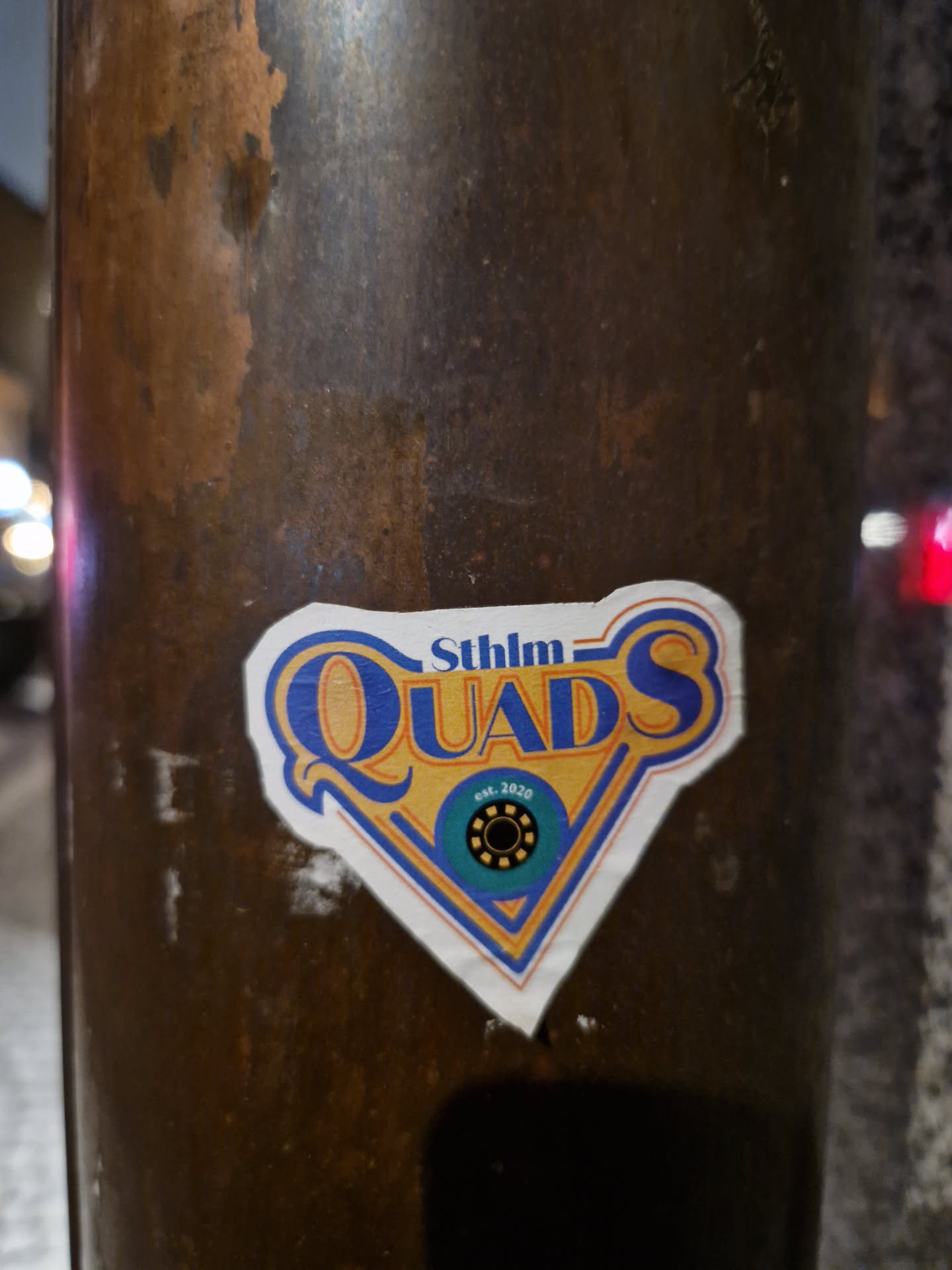 A sticker featuring the logo for Stockholm Quads, a rollerblading group. The logo is a stylized diamond shape with the text 'Sthlm QUADS' in blue and yellow, and a smaller circular logo inside. The sticker is affixed to a dark brown metal pole.