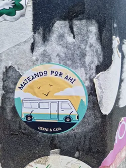 Round sticker with illustration of a van in a landscape, with text in Spanish that says "Mateando por ahí" which translates to "Having mate out there", and the names Herni & Cata at the bottom. 