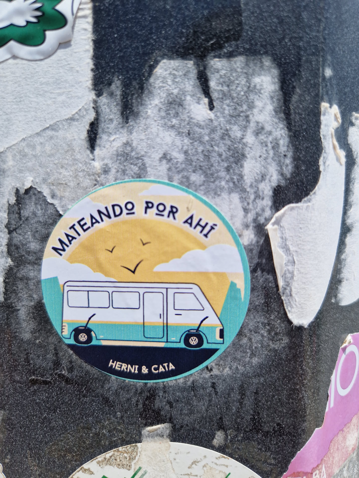 Round sticker with illustration of a van in a landscape, with text in Spanish that says "Mateando por ahí" which translates to "Having mate out there", and the names Herni & Cata at the bottom. 