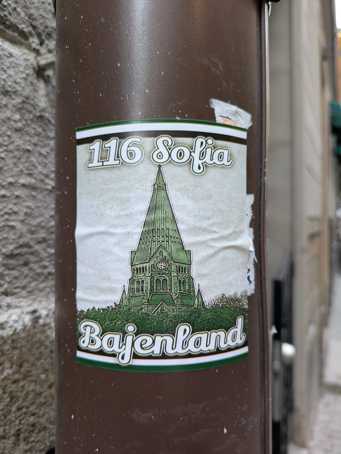 A sticker featuring an illustration of a church with the address "116 Sofia" and the name "Bajenland" written below it. The style is vintage-inspired, with a faded color palette and a slightly worn look.