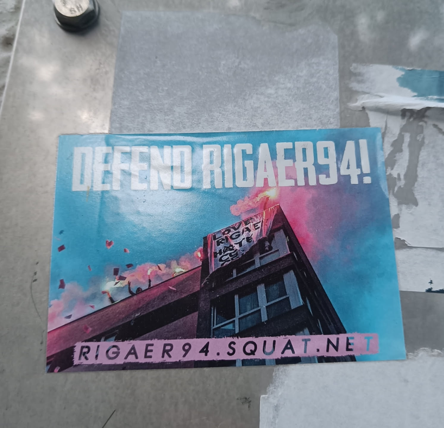 Sticker supporting Rigaer94 squat. The sticker shows a building with a banner and flares. The text on the banner is partially visible and says something about love and hate.  The website address rigaer94.squat.net is shown at the bottom.