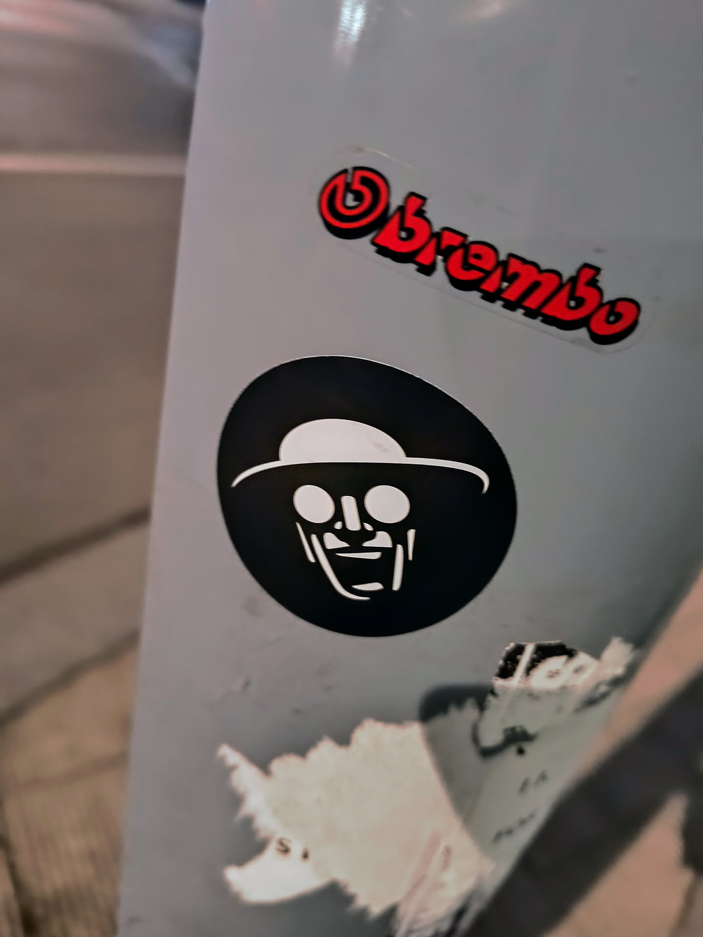 A sticker featuring a black circle with the minimalist white outline of a smiling man wearing a wide-brimmed hat. Above it there is a red Brembo sticker.