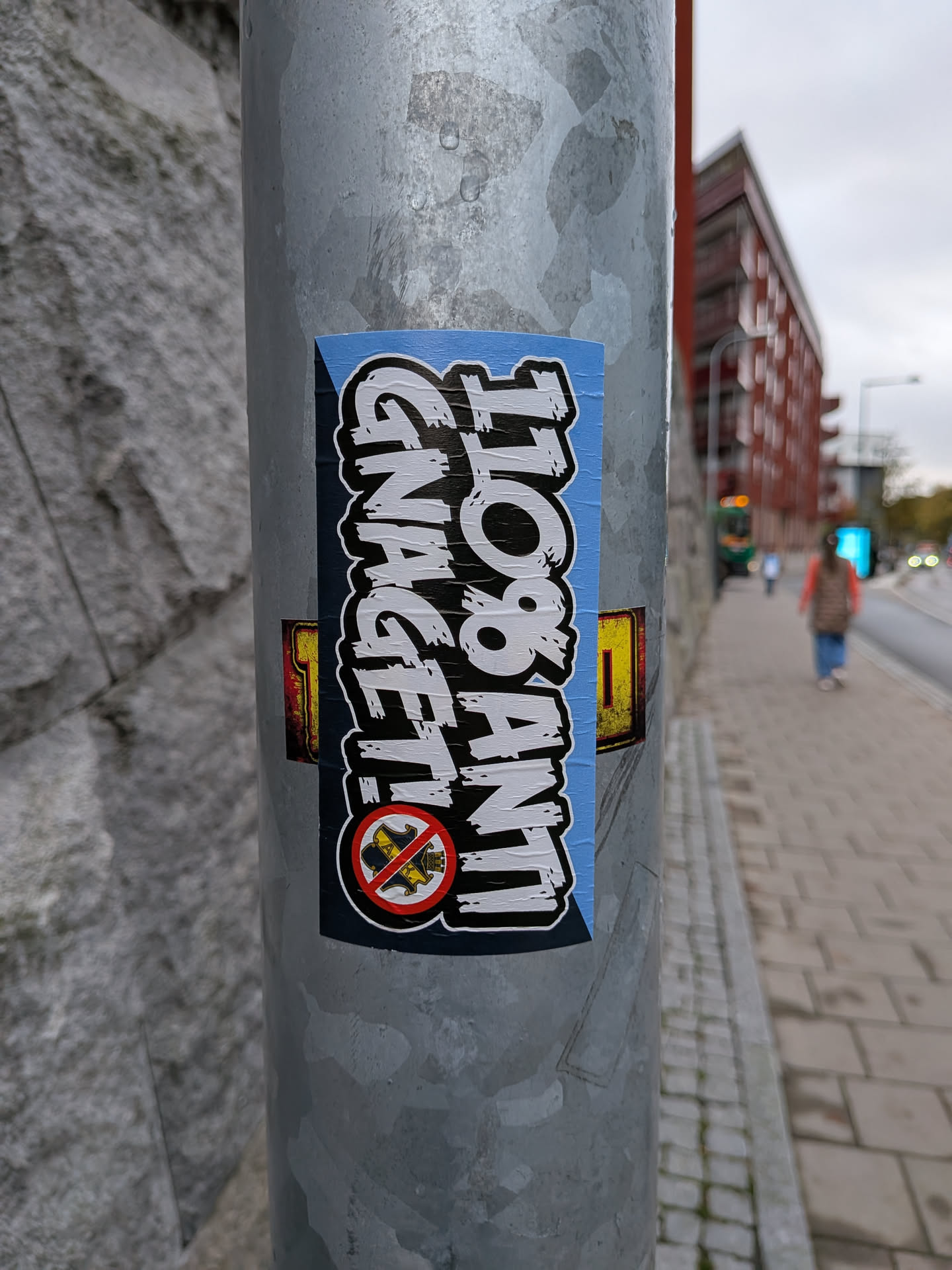 A vertical rectangular sticker adhered to a galvanized metal pole. It features stylized, distressed white text with heavy black borders reading '110% ANTI' and 'GNAGET!' arranged vertically. The background consists of light blue and dark blue sections divided diagonally. In the lower left corner, there is a prohibition sign with a red circle and slash crossing out the AIK football club crest.