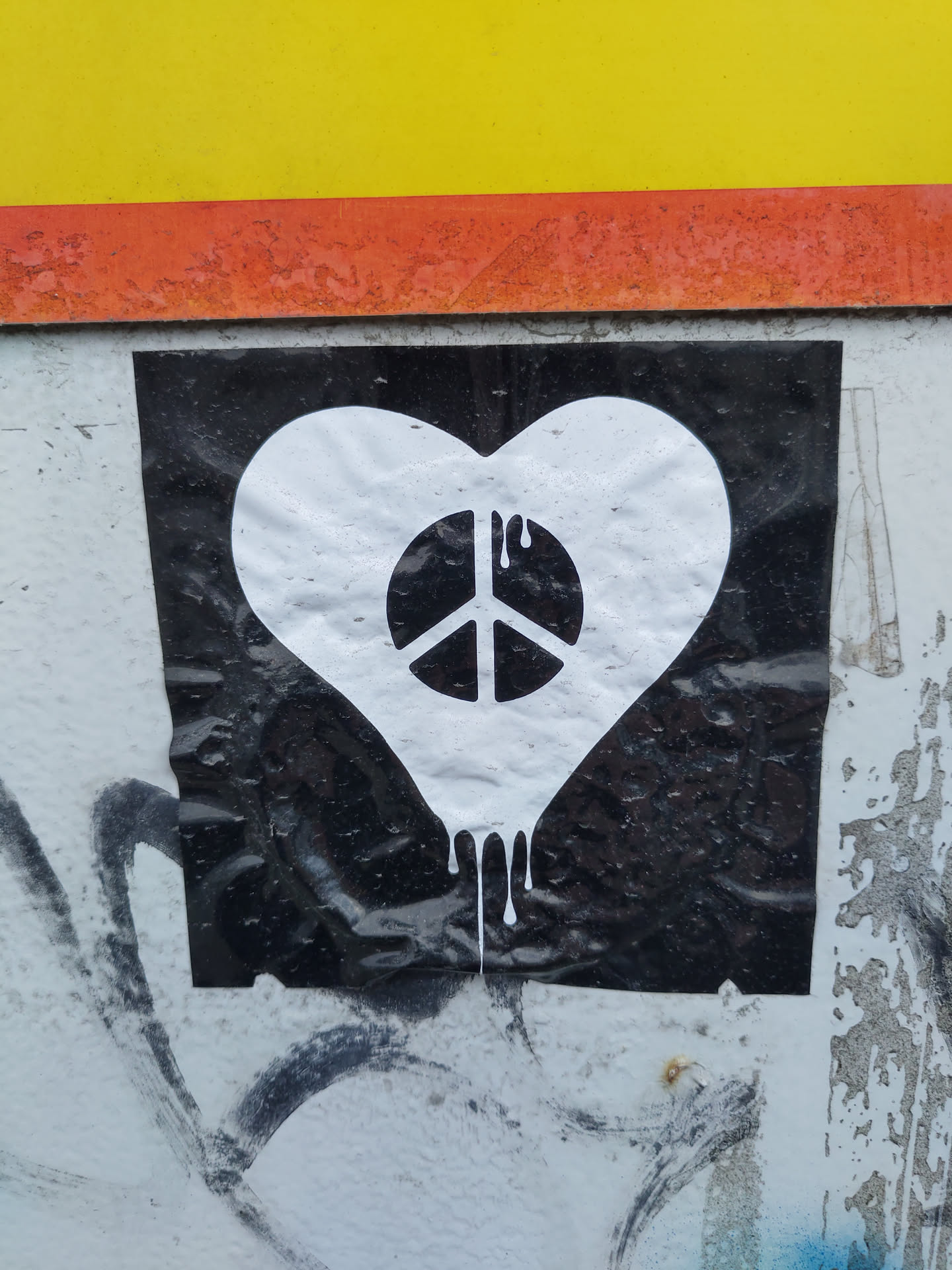 A black and white sticker on a wall shows a white heart shape with a peace symbol inside.  The heart appears to be dripping. The sticker is affixed to a wall with some existing graffiti.