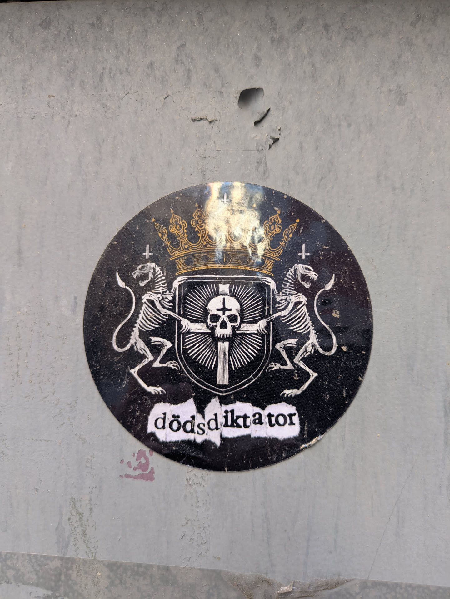 The sticker is a round, black and white design with a golden crown at the top. Below the crown is a heraldic shield with a cross and a skull. Two skeletal lions flank the shield, each holding a sword. At the bottom, the word "dödsdiktator" is written in a rough, stylized font.