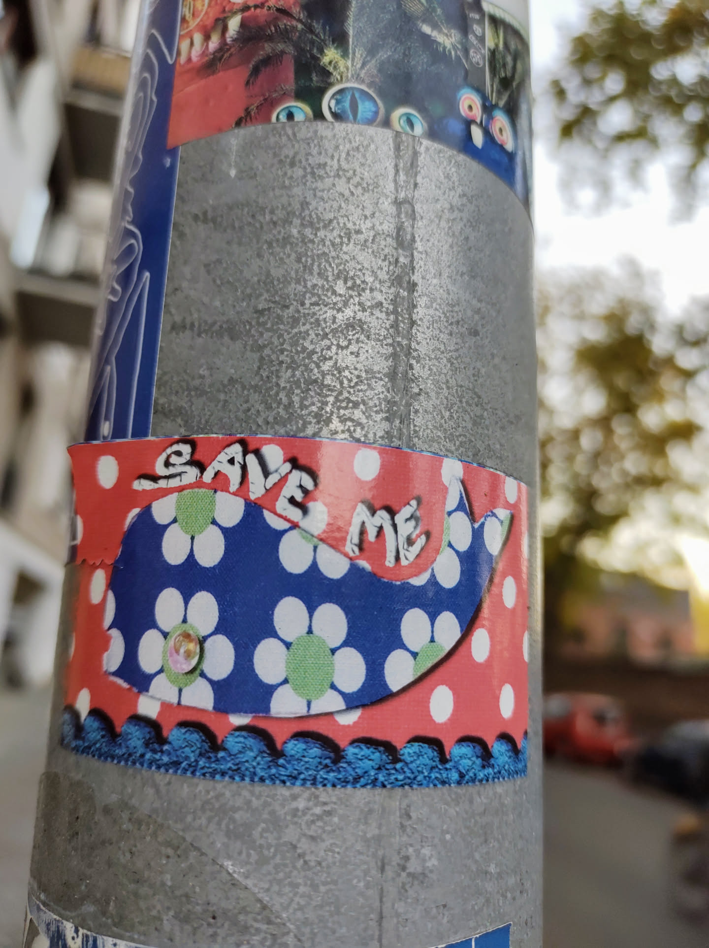 A charming sticker featuring a whale with a floral pattern and the words "SAVE ME" written on it. The sticker is predominantly blue, white, and red, with a scalloped edge. It's affixed to a gray metal pole.