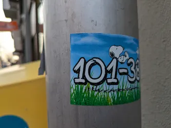 A sticker featuring Snoopy lying down, set against a backdrop of a blue sky and green grass with barbed wire. The numbers 101-38 are prominently displayed.
